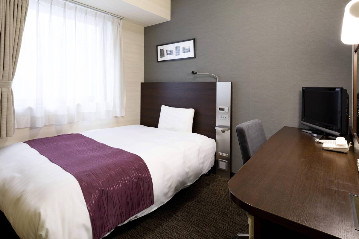 A hotel room with a large bed, a desk with a computer, a TV, and a window with curtains.
