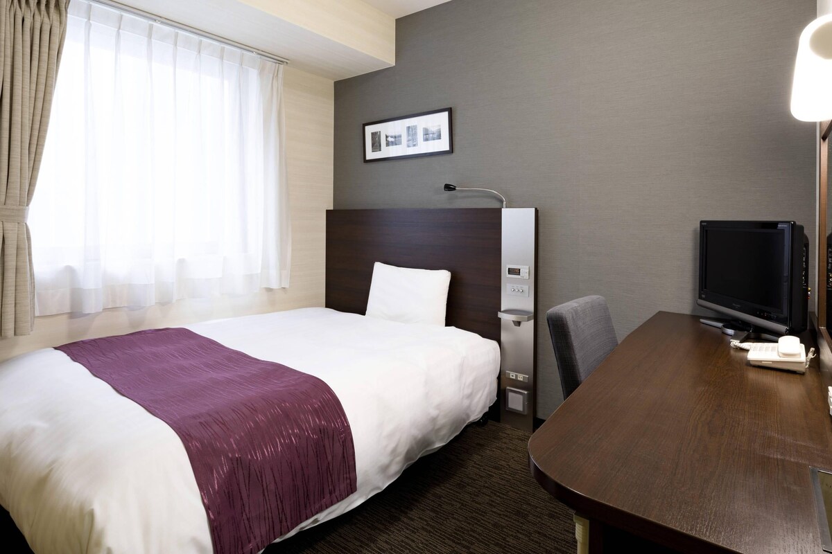 A hotel room with a large bed, a desk with a computer, a TV, and a window with curtains.
