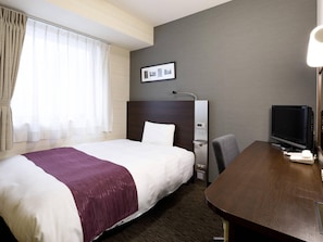 A hotel room with a large bed, a desk with a computer, a TV, and a window with curtains.
