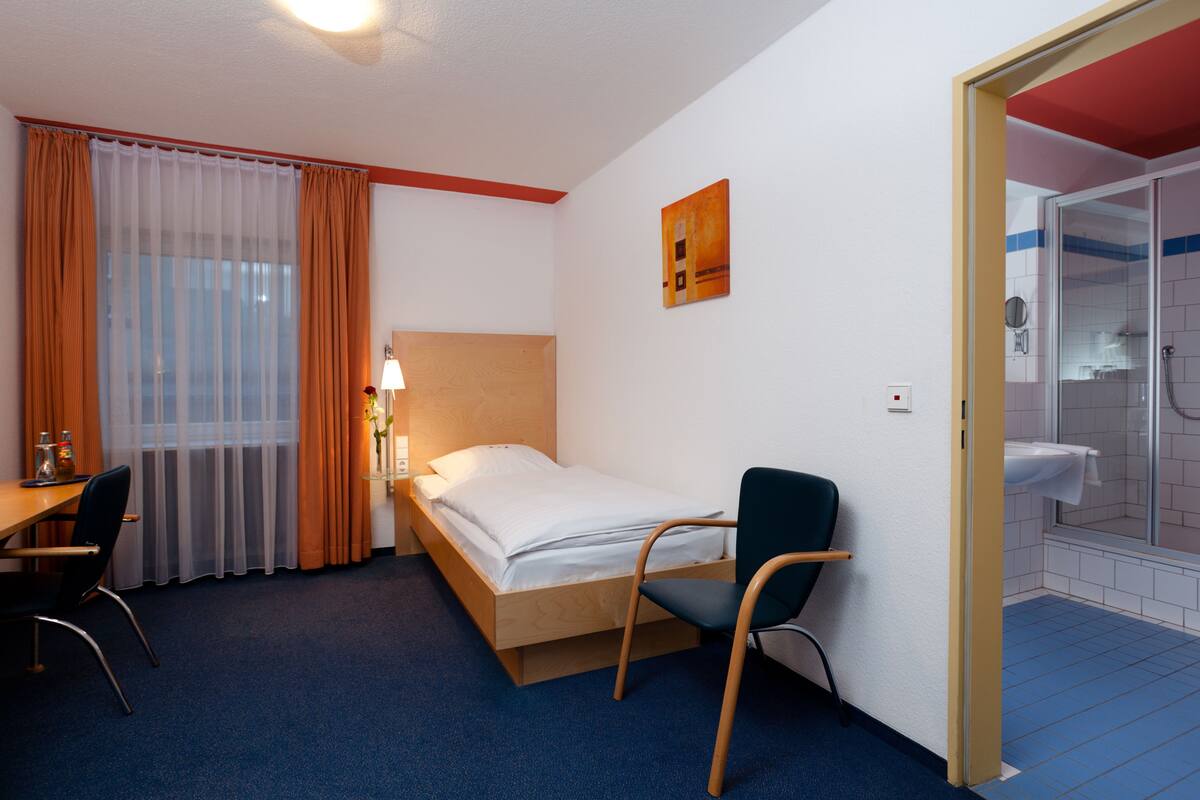 Une chambre d’hôtel comprenant un lit, un bureau, une chaise, une lampe, une fenêtre avec des rideaux et un espace douche.