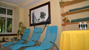 A room with three blue lounge chairs, a yellow table with two bottles, a hammock decoration, and shelves with plants and bottles.