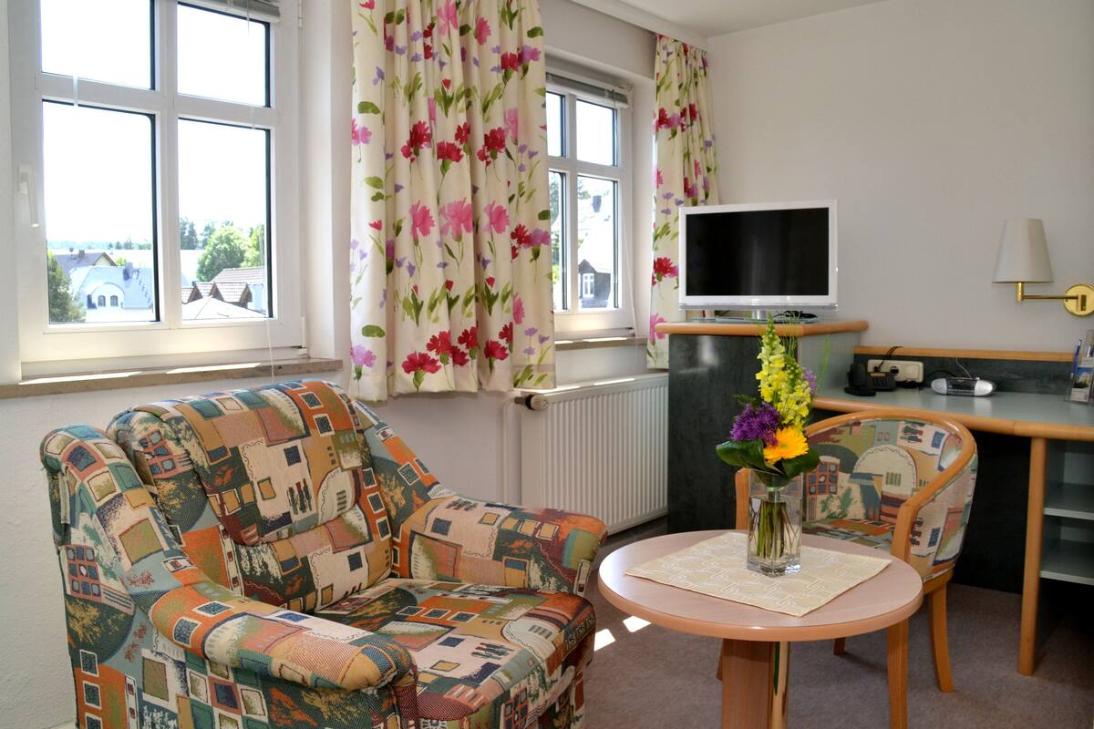 A cozy room with a patterned armchair, a small round table with a vase of flowers, a television on a stand, and a window with floral curtains.