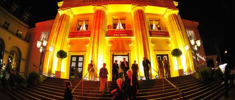 El exterior del Teatro König Albert con escalones que conducen a la entrada, iluminado por una cálida luz.
