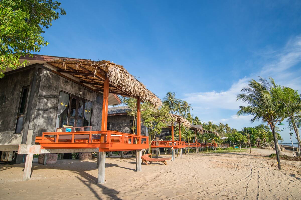 Un resort frente a la playa con cabañas de techo de paja, sillas de madera y palmeras.