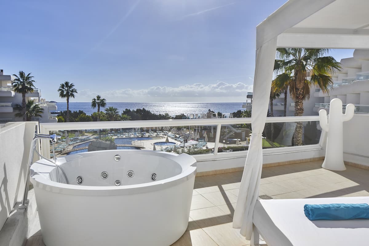Un balcón con bañera de hidromasaje, cama y vista al mar y edificios del complejo.
