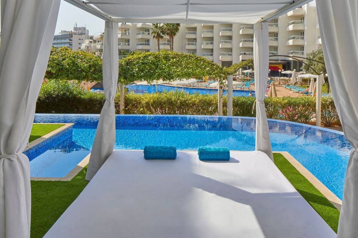 Zona junto a la piscina con una toldo blanco, dos toallas y vistas a una piscina grande y edificios al fondo.