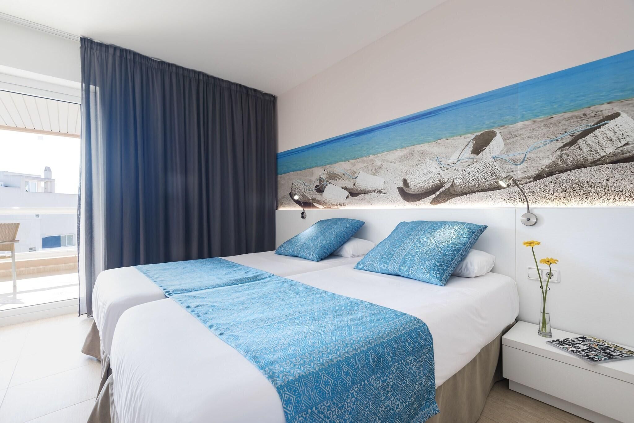 A double bed with a blue patterned bedspread, a nightstand with a vase of flowers, and a painting of a beach scene on the wall.