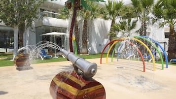 Een waterfontein met een brede, cilindrische basis en een metalen tuit, omringd door palmbomen en een modern gebouw op de achtergrond.