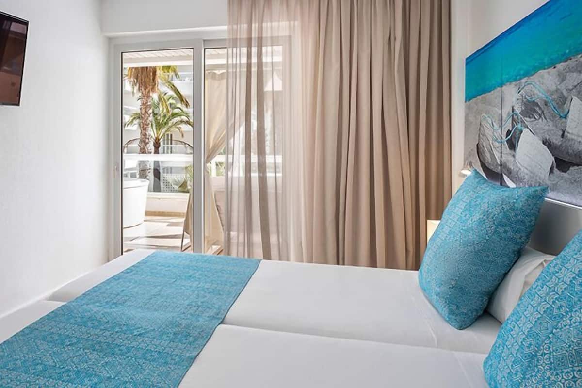 Habitación de hotel con cama, televisor montado en la pared y balcón con vista a palmeras.