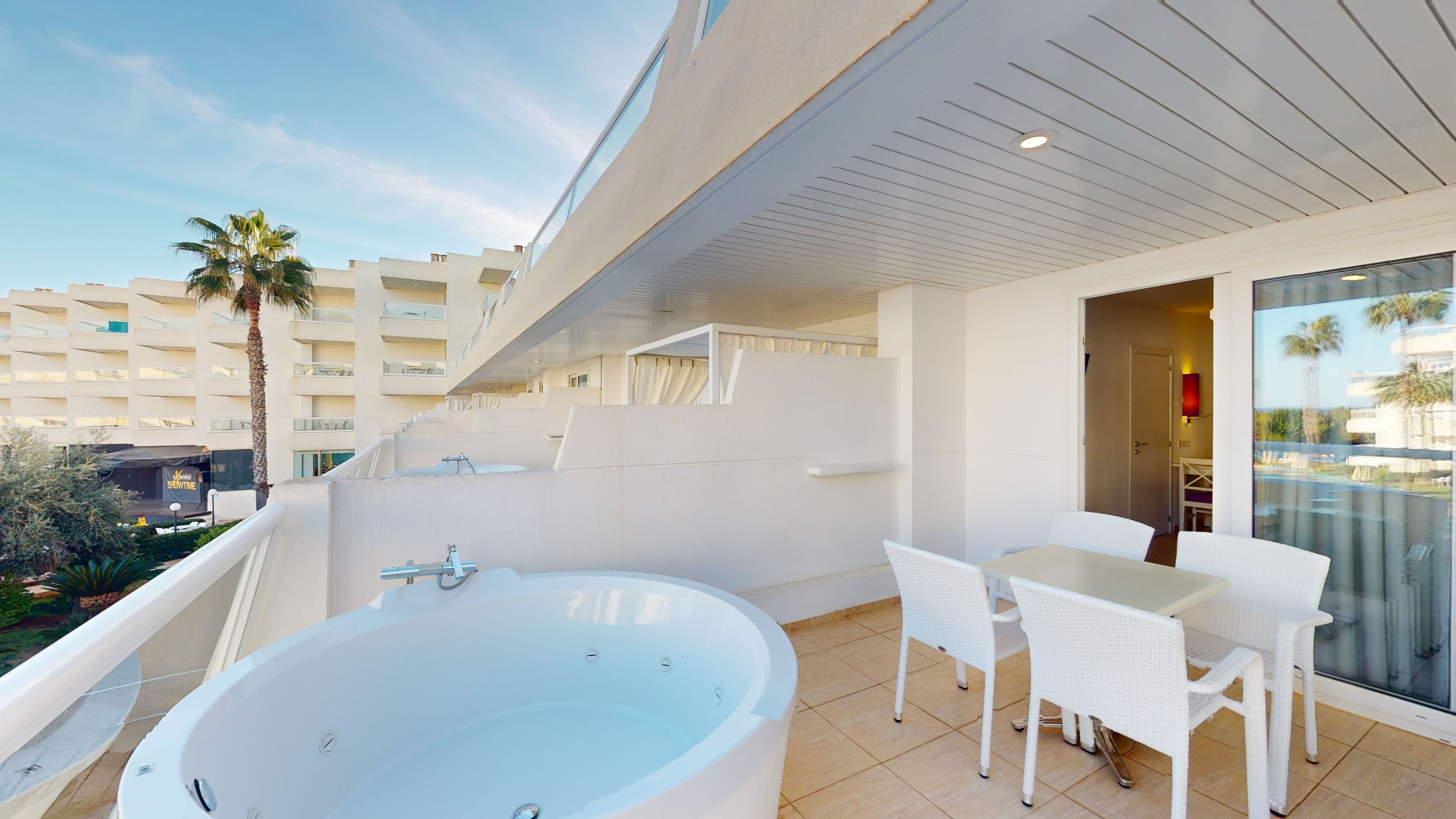 A balcony with a hot tub, white wicker chairs, and a view of a hotel building.