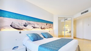 A bedroom with a bed, a nightstand, a desk, and a view of the beach through a window. - Aparthotel Tropic Garden (Santa Eulalia del Rio)