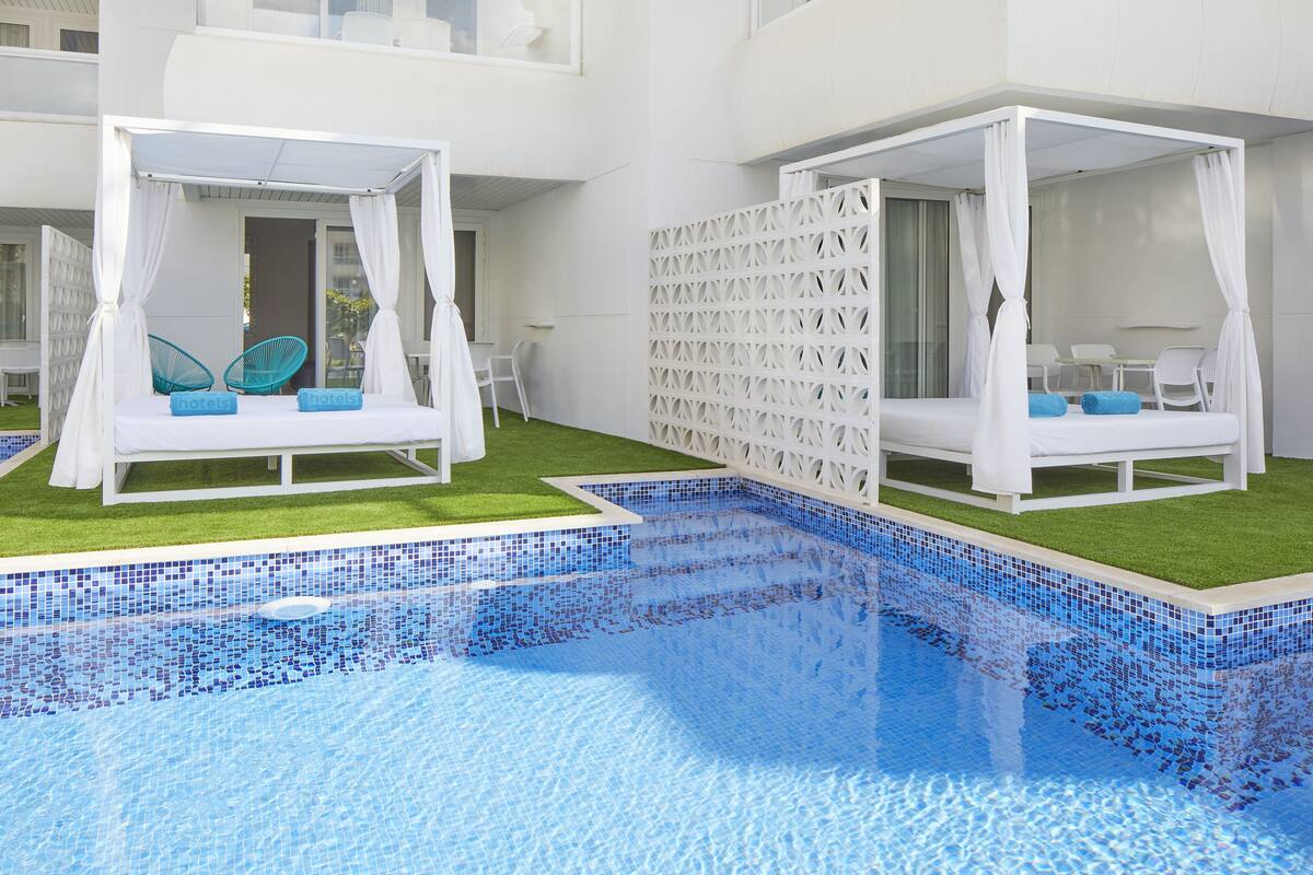 Un área de piscina con dos camas y sillas blancas con toldos, rodeada por una cerca blanca y un edificio con balcones.