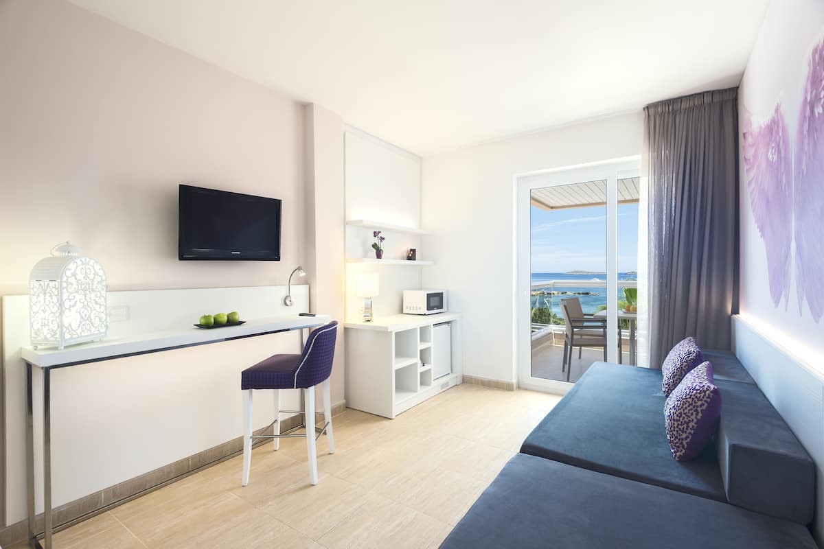 Una habitación de hotel moderna con kitchenette, un televisor de pantalla plana, un sofá con cojines decorativos y un ventanal con vistas al mar.