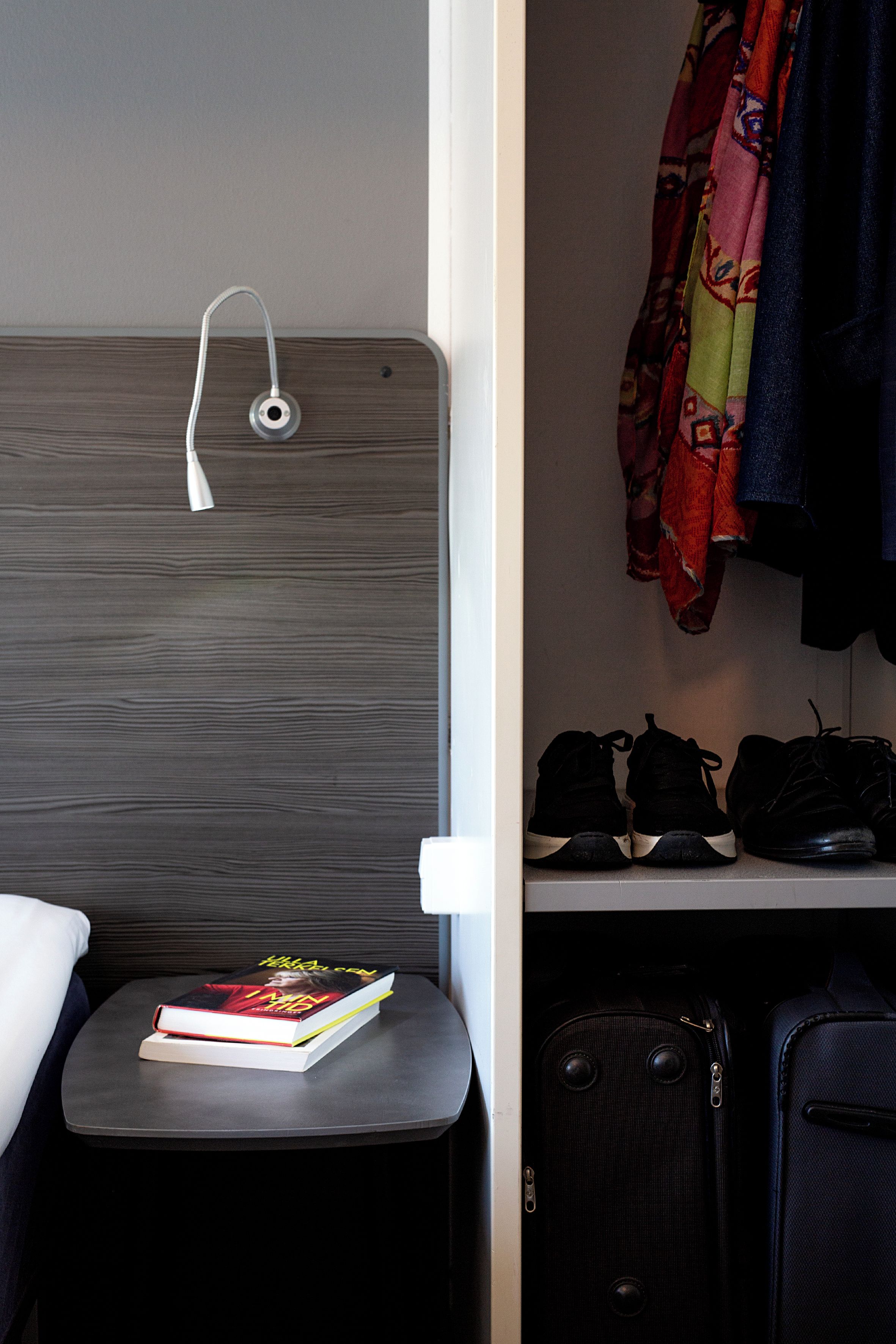 A hotel room with a bed, a bedside table with a book, a suitcase, and a shelf with shoes and a hanging garment.