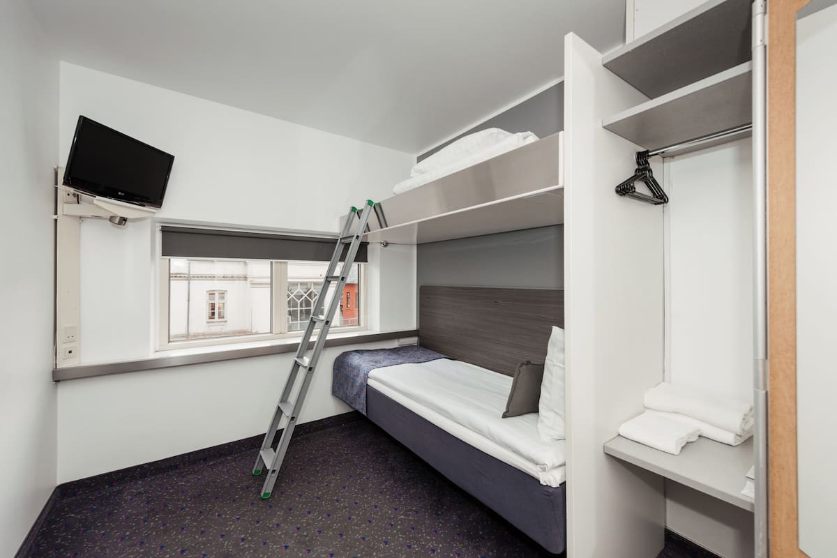 Een kleine hotelkamer met stapelbedden, een aan de muur bevestigde televisie, een raam met uitzicht op gebouwen en een ladder voor toegang.