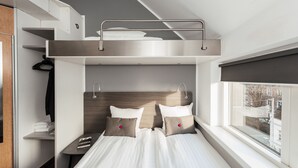 A compact hotel room with a bunk bed, a small desk with a chair, a window with a view of buildings, and a wardrobe.