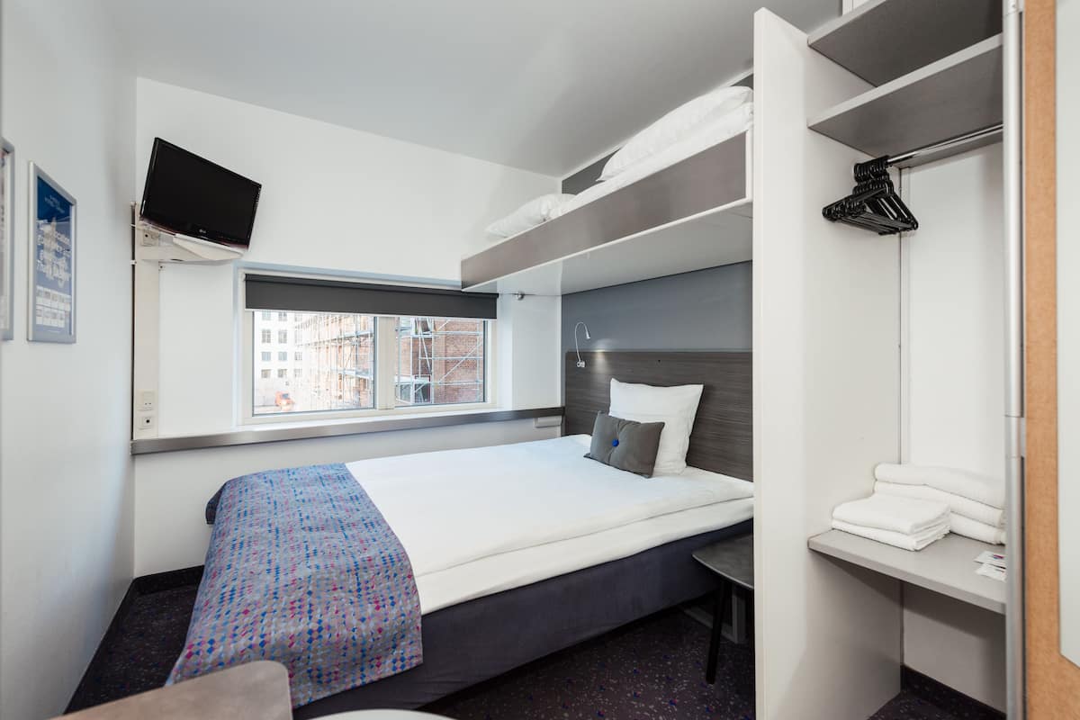 Hotelkamer met een bed, een aan de muur bevestigde televisie, een raam met uitzicht op gebouwen en een inbouwkleerkast met hangende kleding.
