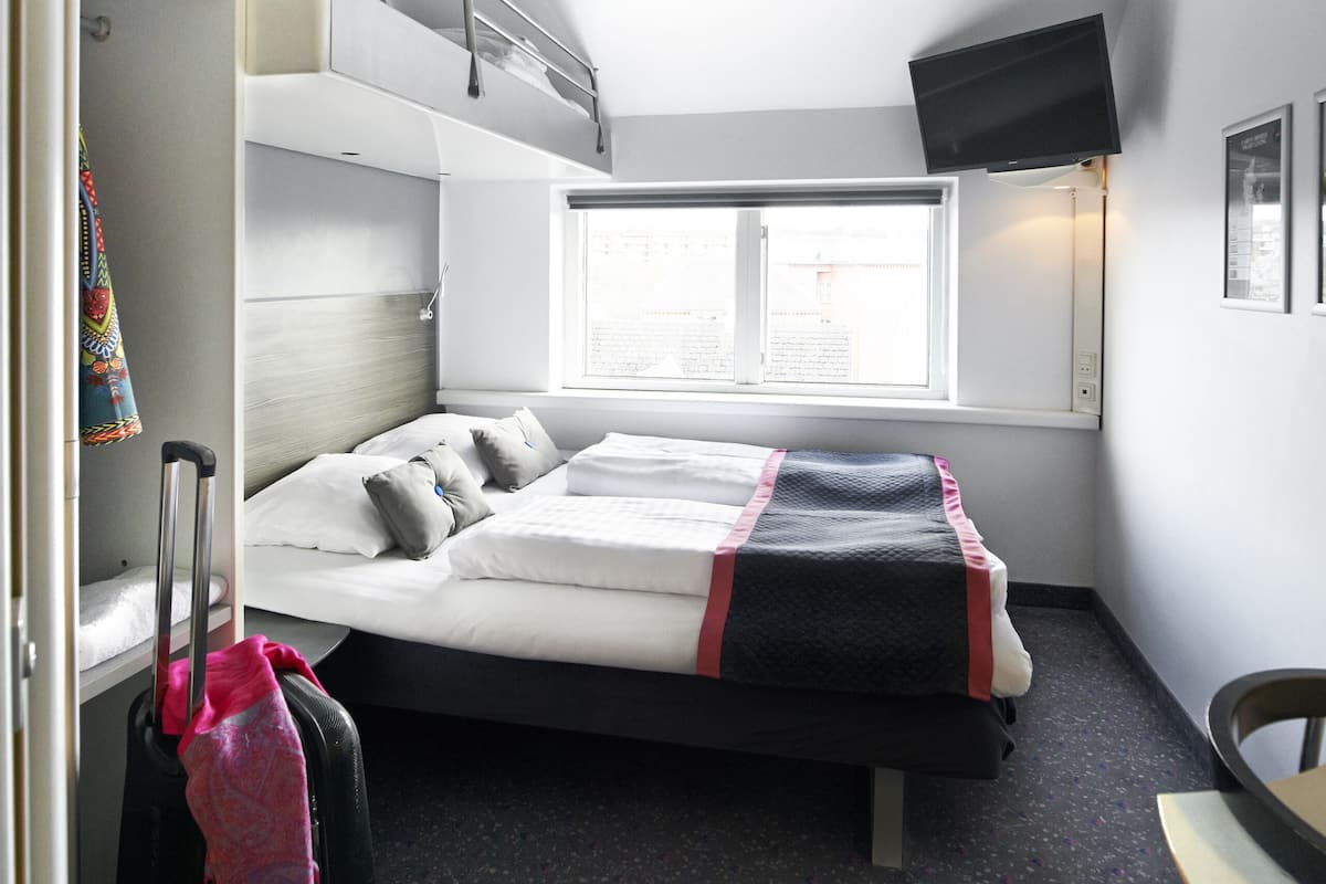 Een kleine hotelkamer met een stapelbed, een bureau, een stoel, een koffer en een raam met uitzicht.