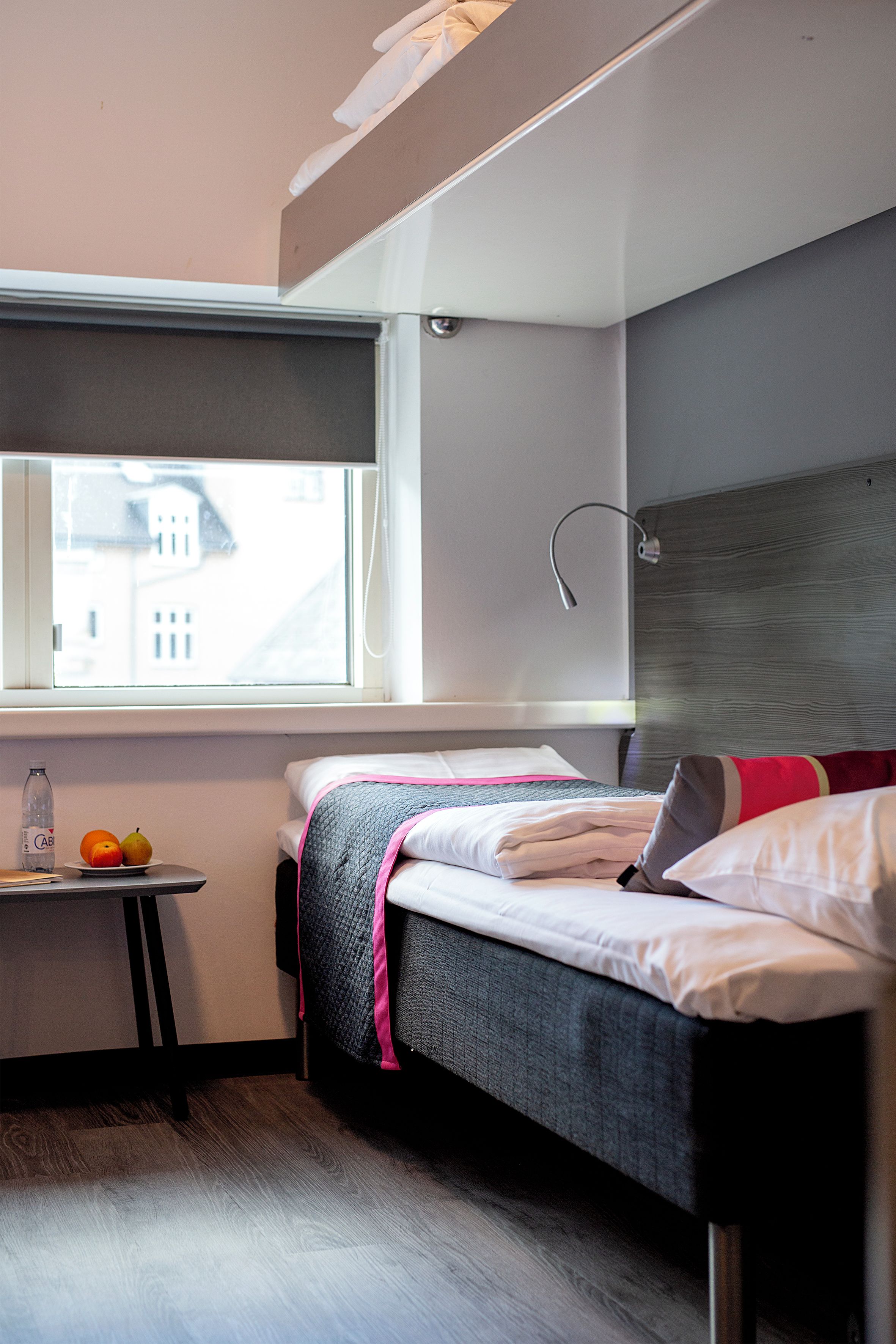 Et lille soveværelse med en enkeltseng, et lille bord med frugt, et vindue med udsigt til bygninger og en sengegavl med en lampe.