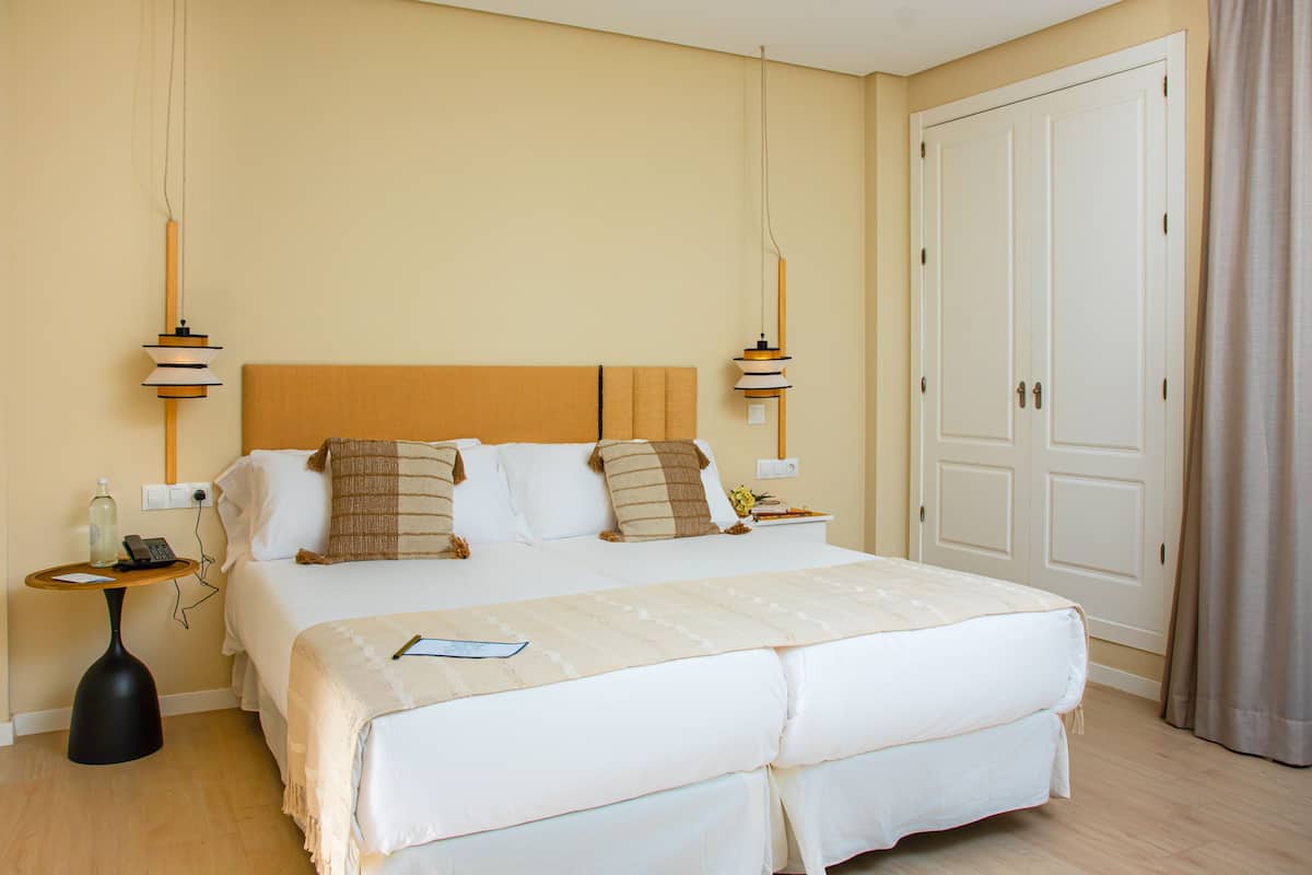 A neatly made bed with a white comforter and multiple pillows, a bedside table with a lamp, and a door with a curtain.