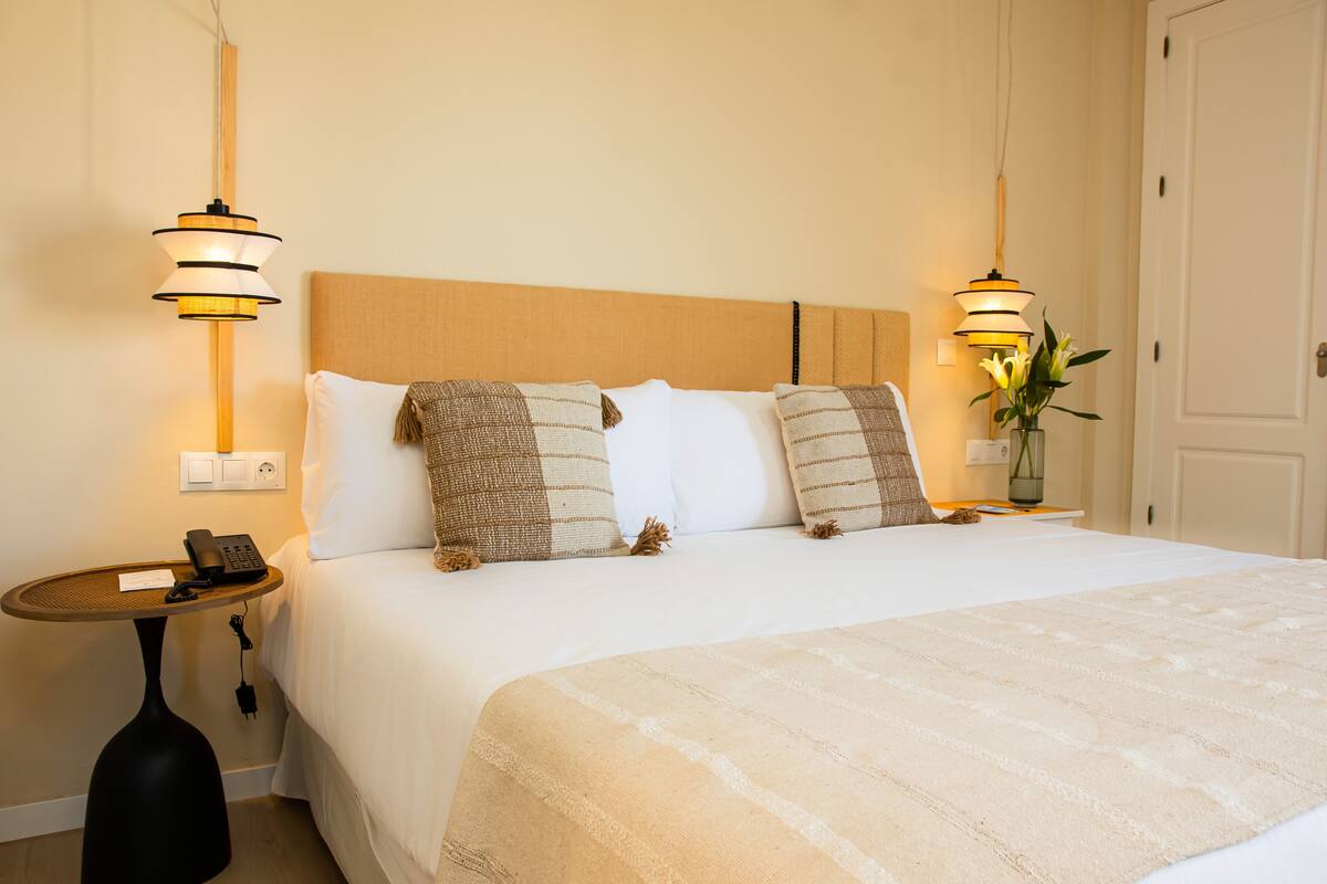A neatly made bed with a striped blanket, two bedside lamps, a bedside table with a phone, and a vase with flowers.