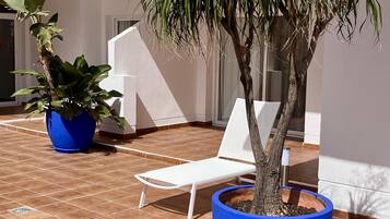 Un patio con una silla lounge blanca, dos plantas grandes en macetas y suelo de baldosas.