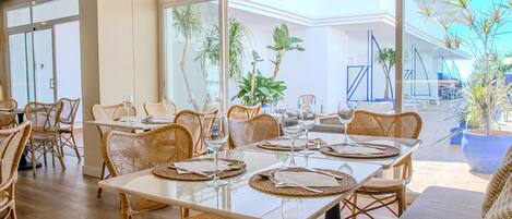 Un vaste espace dédié à la salle à manger, avec des chaises et des tables en osier, de grandes fenêtres et des plantes en pot.