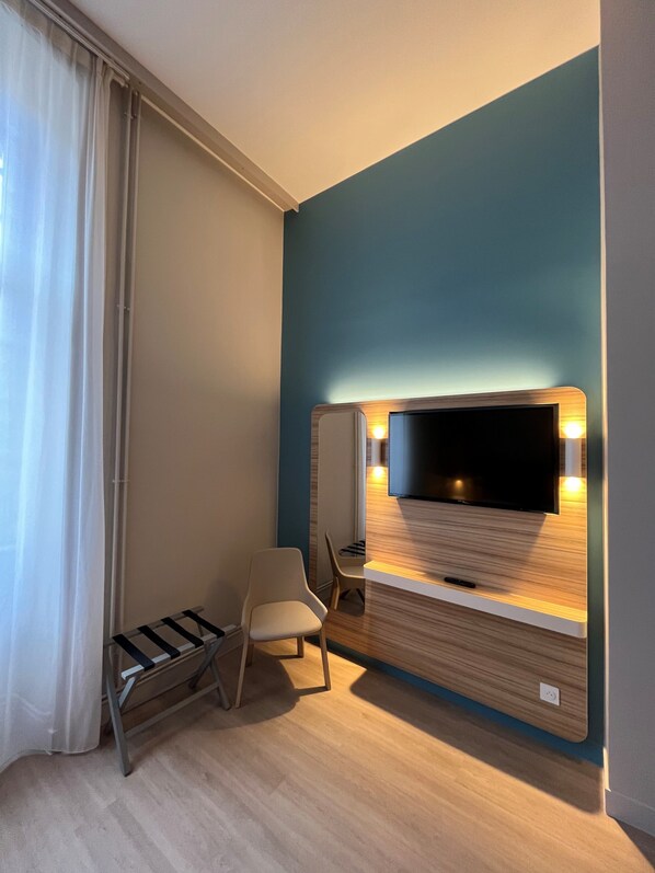 A modern hotel room with a flat-screen TV mounted on a wooden panel wall, a small seating area with a chair, and a window with sheer curtains.