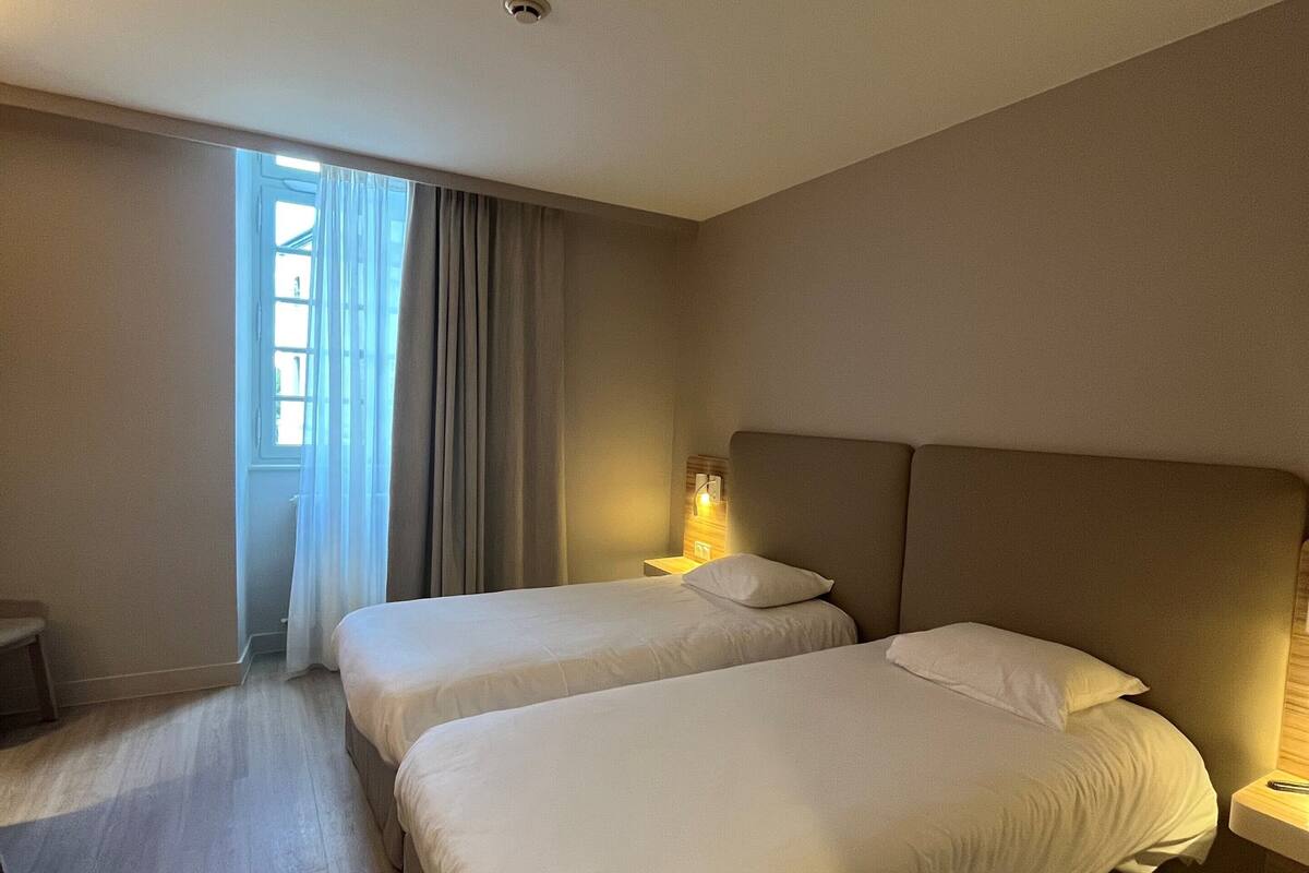A hotel room with two beds, a bedside table, a lamp, and a window with curtains.