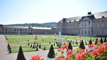 A large, historic building with a symmetrical garden layout, including geometrically shaped hedges and a central monument.