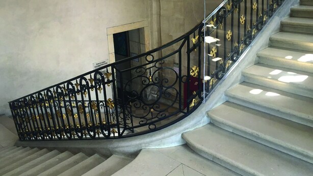 Un magnifique escalier avec des rampes noires richement décorées, un grand lustre et des éléments architecturaux classiques.