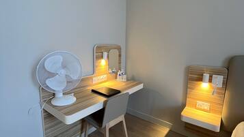 A small, well-lit room with a wooden desk, a chair, a standing fan, and a bedside table with a lamp.