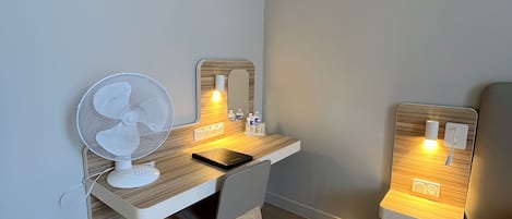A small, well-lit room with a wooden desk, a chair, a standing fan, and a bedside table with a lamp.