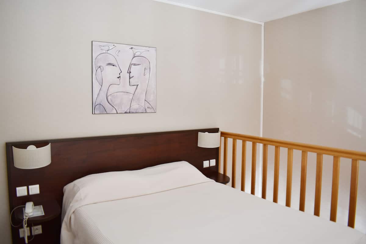 A hotel room with a wooden headboard, a white bedspread, a wall-mounted artwork depicting two stylized faces, and a bedside table with a lamp and a phone.