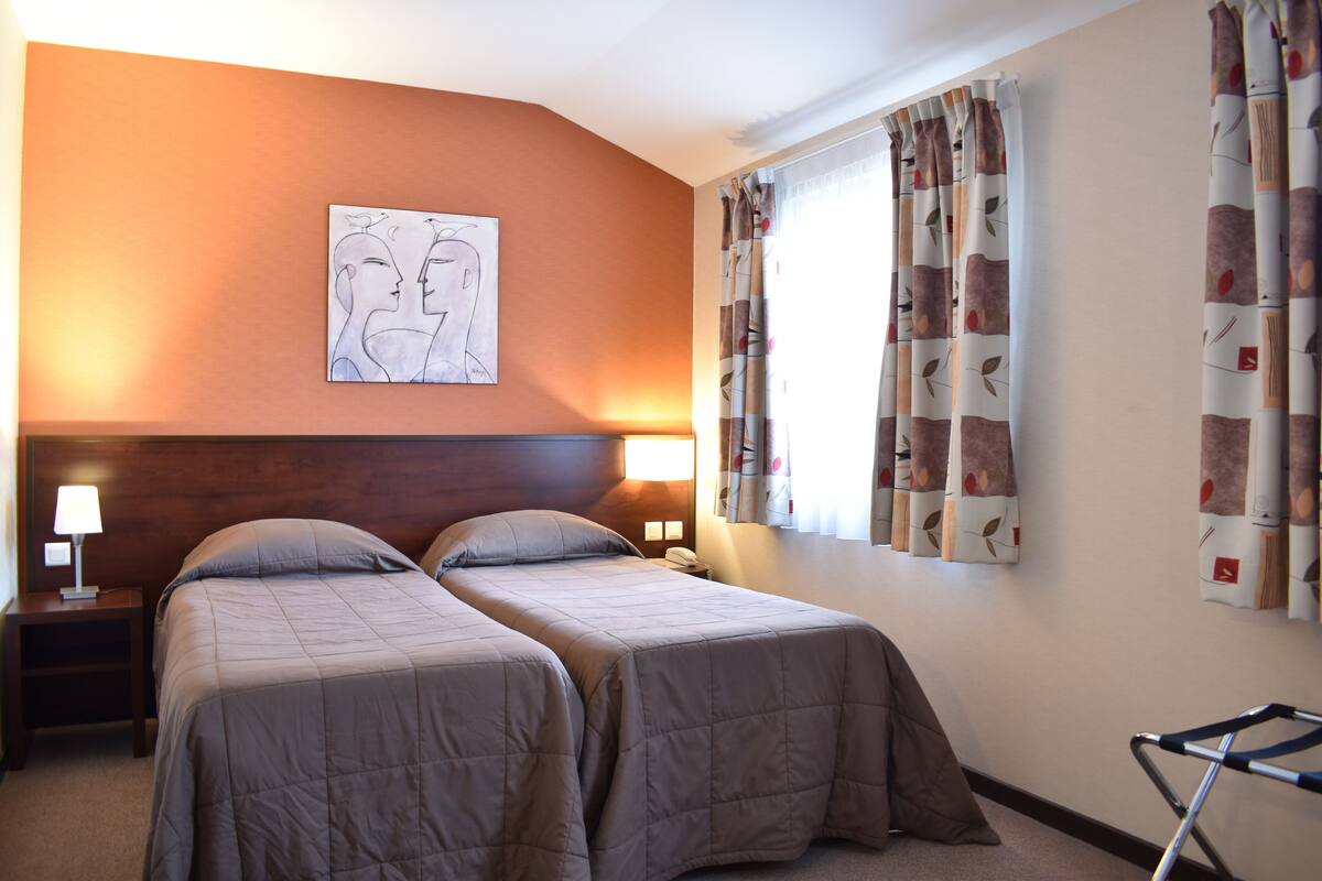 A hotel room with two beds, a headboard, a nightstand with a lamp, a wall-mounted artwork, and a window with curtains.