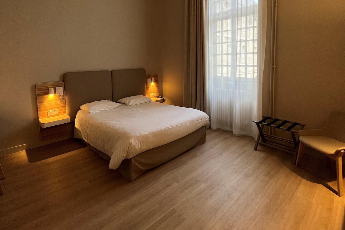 A bedroom with a large bed, bedside lamps, a bench, and a window with curtains.