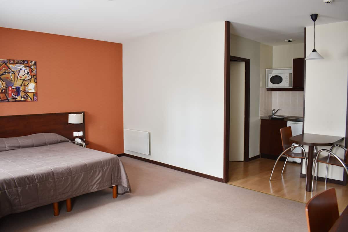 A hotel room with a bed, a desk, and a chair. There is a painting on the wall and a microwave in the kitchen area.