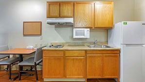 A compact kitchen with wooden cabinets, a microwave, a refrigerator, a sink, and a dining table with chairs. - Extended Stay America Select Suites - Phoenix - West (Phoenix)