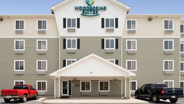 Un hotel a più piani con ingresso centrale, condizionatori d'aria e un'area parcheggio con due veicoli.