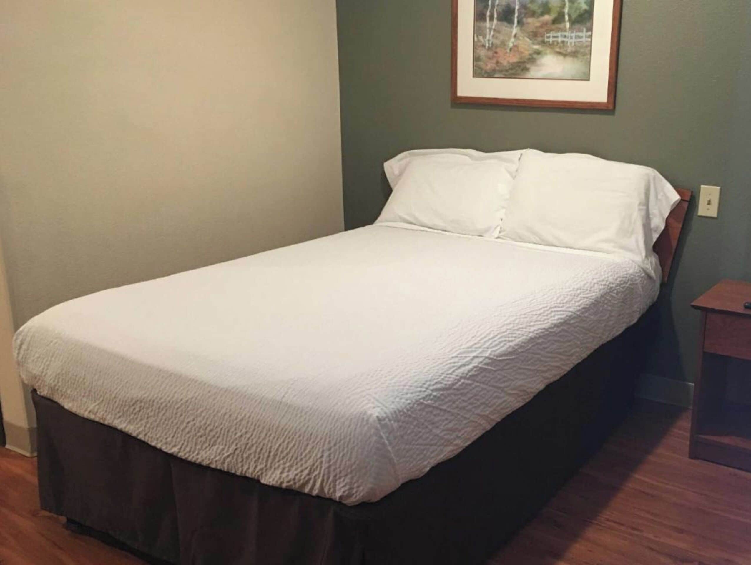 Una cama individual con colchón y almohadas blancos, cabecera de madera y un cuadro en la pared.
