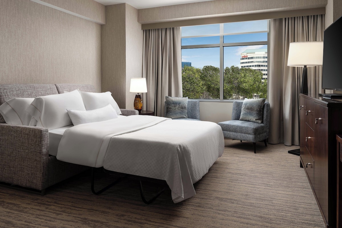 A hotel room with a large bed, a seating area, a TV, and a window with a view of a building and trees.