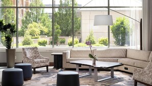 A modern hotel lobby with a large window, a sofa, ottomans, a coffee table, and a potted plant. - The Westin Reston Heights (Reston)
