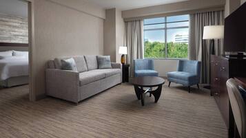 A hotel room with a sofa, armchair, small table, lamp, TV, and a view of greenery outside.