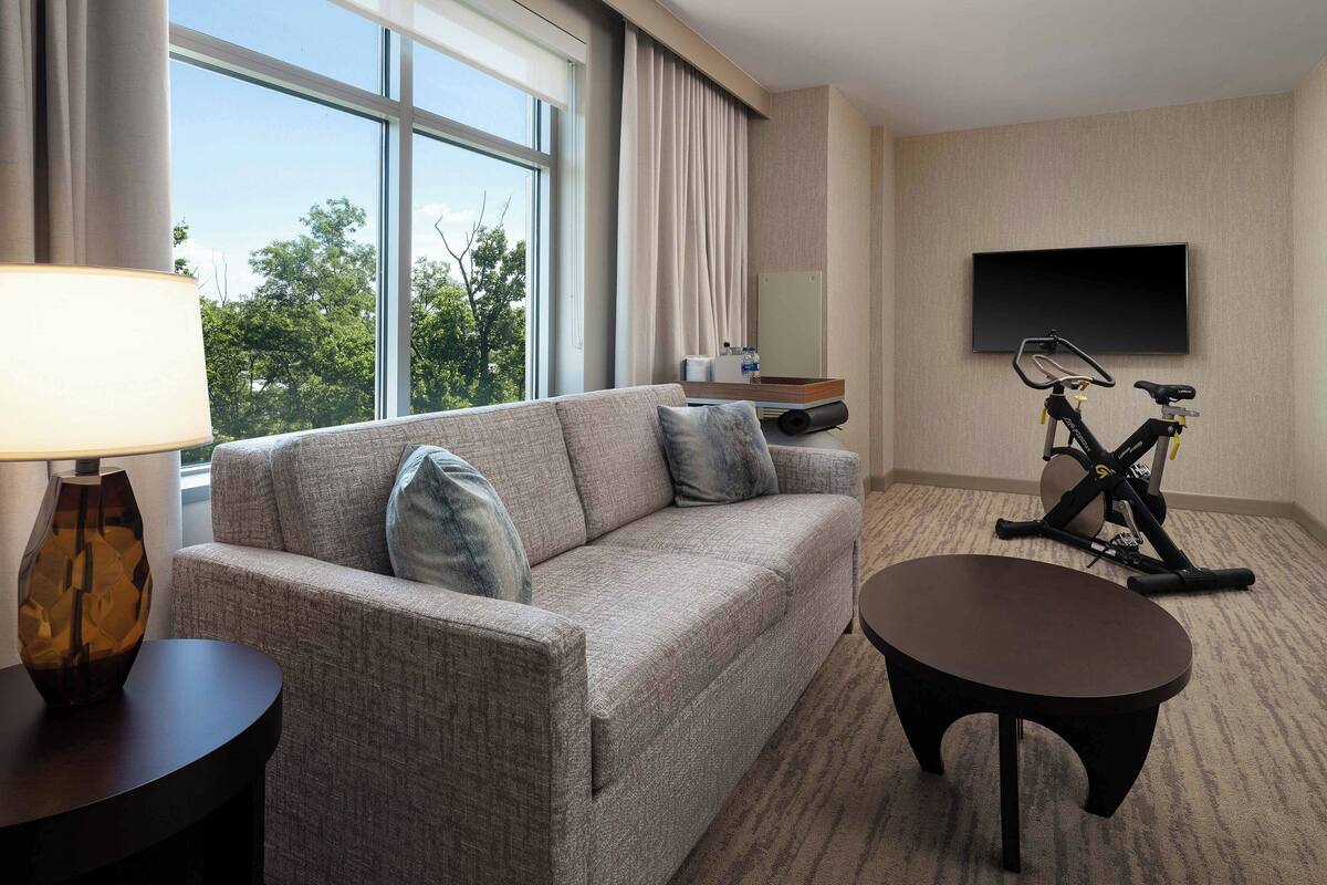 A hotel room with a sofa, a small table, a TV mounted on the wall, and an exercise bike.