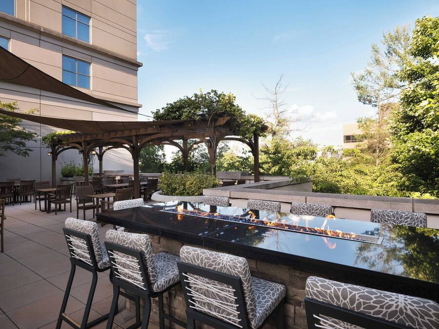 An outdoor bar area with a long table and multiple chairs, surrounded by greenery and a pergola.