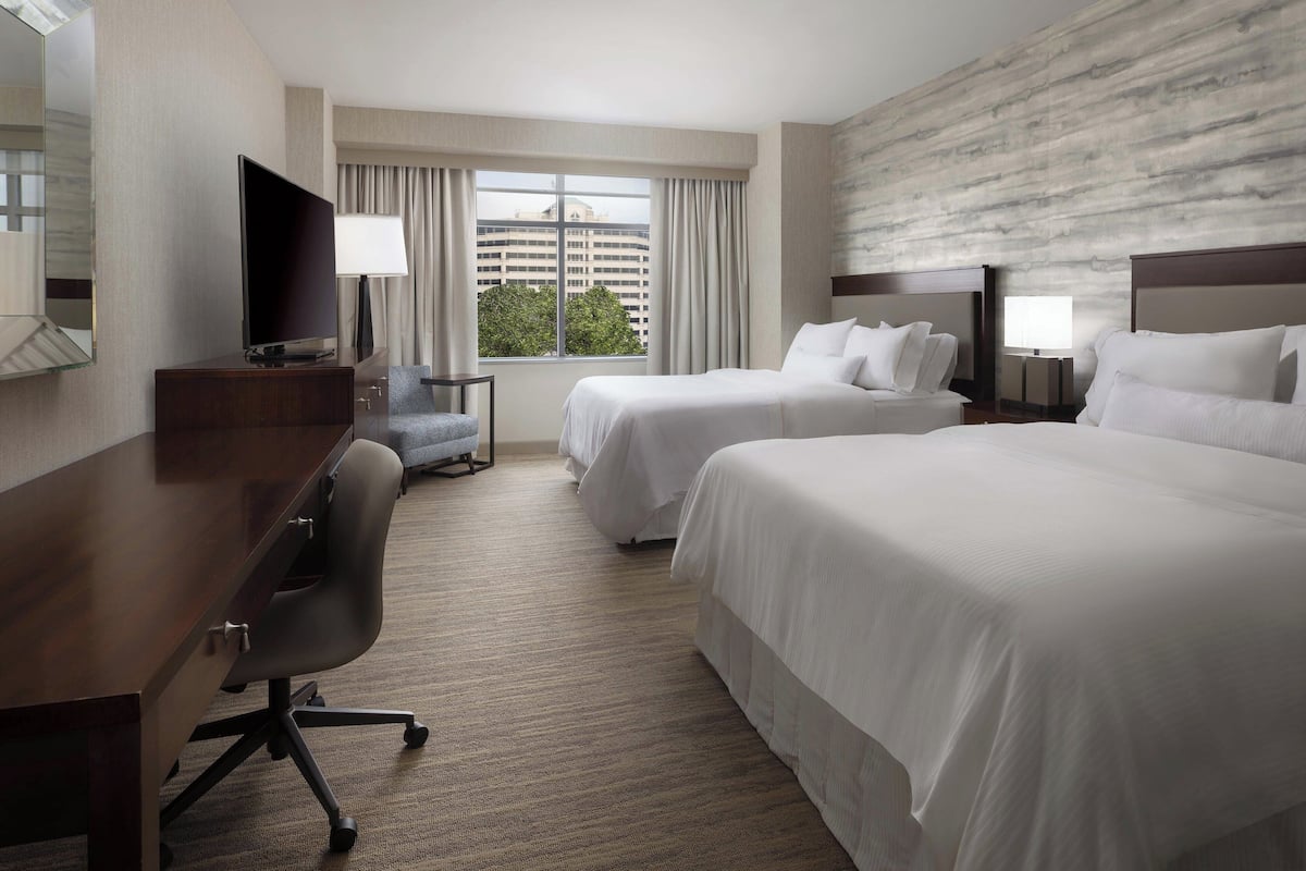 A hotel room with two beds, a desk, a chair, a TV, and a large window with curtains.