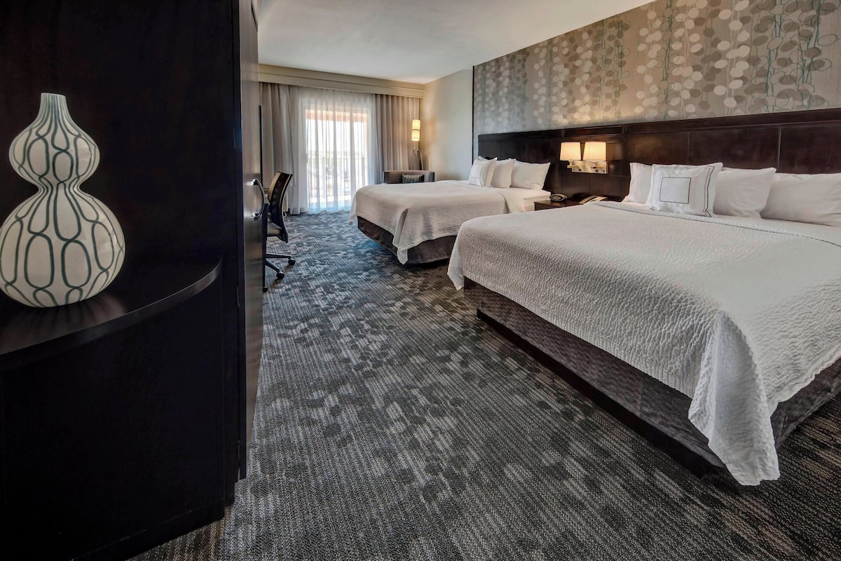 A hotel room with two beds, a dark wood headboard, a patterned wall, and a large vase on a dark console table.