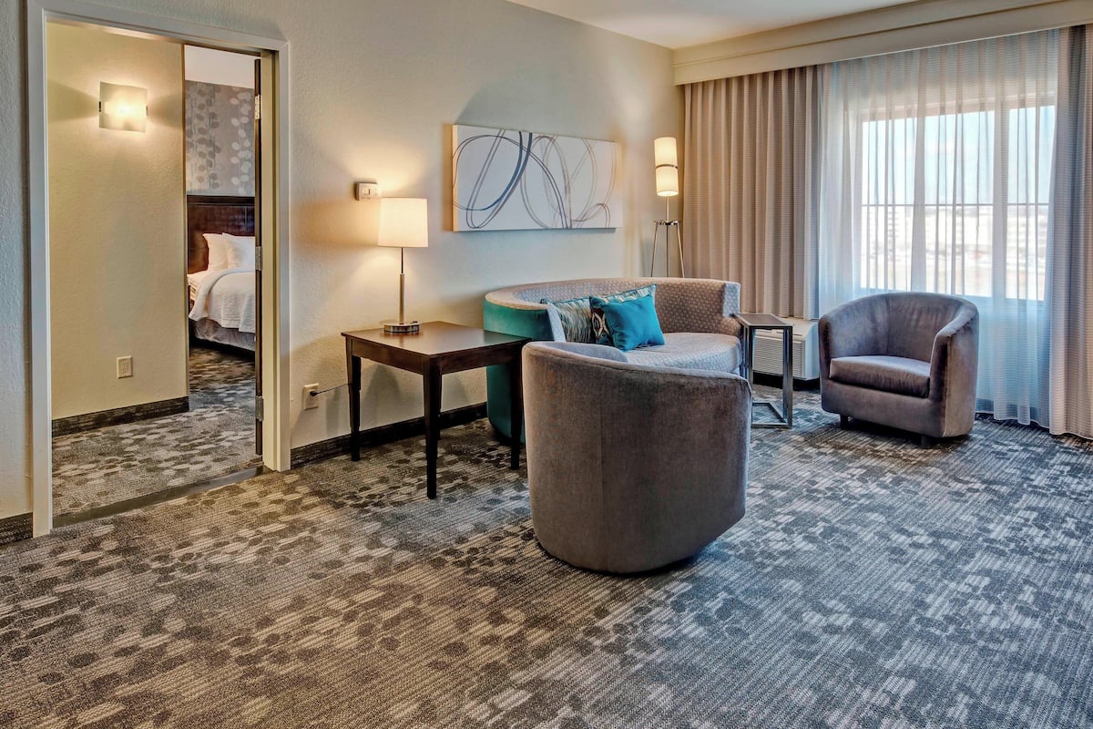 A hotel room with a carpeted floor, a sofa, a chair, a small table, a lamp, a painting on the wall, and a bed in the background.