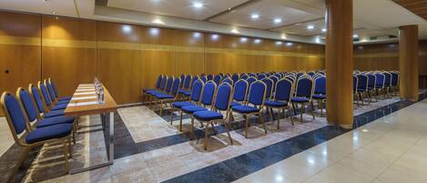 A conference room with rows of blue chairs and a long table, wooden panel walls, and a tiled floor.