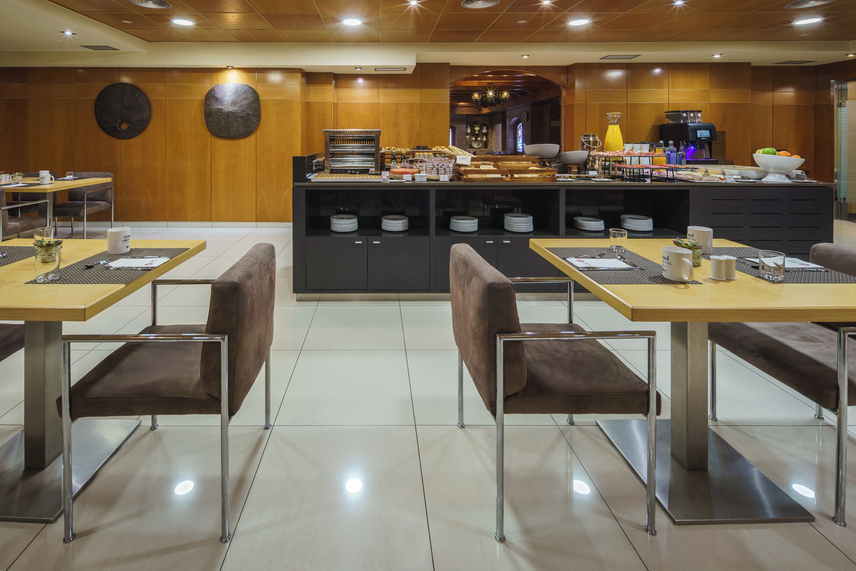 A modern hotel dining area with wooden panel walls, a central buffet, and two tables with chairs.